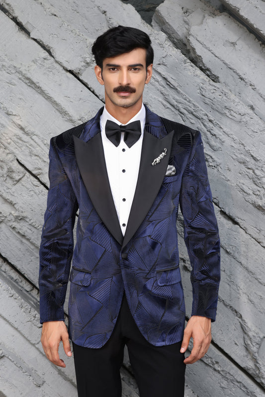 A man wearing a blue patterned velvet tuxedo jacket with black satin lapels, a white dress shirt, and a black bow tie, standing against a gray stone wall.