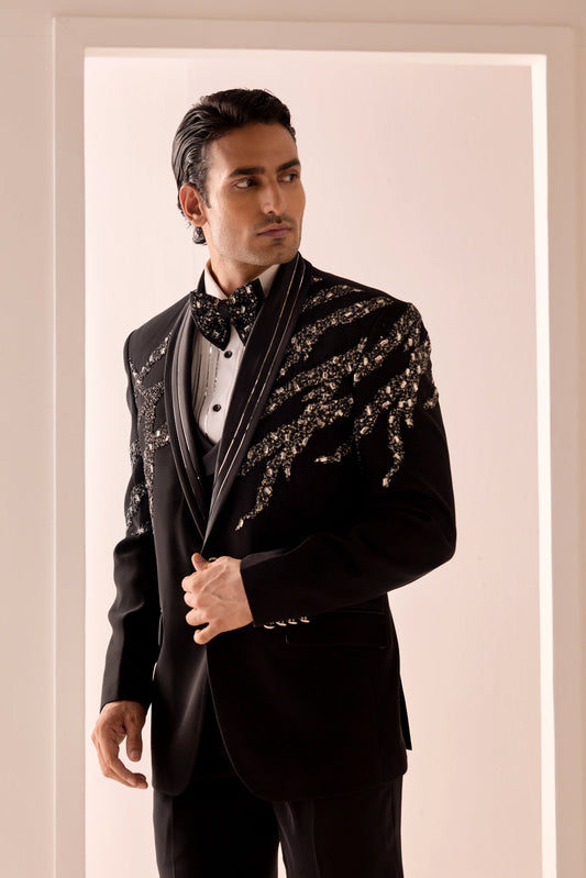 A man wearing a black tuxedo jacket with ornate silver and gold sequin embellishments on the shoulders, paired with a white shirt and black bow tie, standing in a doorway.